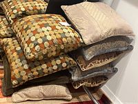 Pile of 9 pillows stacked showing three distinct patterns: beige damask, blue-gray floral, brown solid, and multicolored circles.