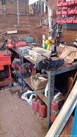 Wide view of metal shelving unit in shed, loaded with two bench vices mounted to shelving and various tools and containers on shelves.