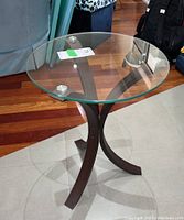Side view of round side table with glass top and dark wooden curved legs on a tile and wood floor.