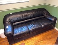 Overall front perspective of black leather-like three-seat sofa placed in a room with green walls and wooden floor.