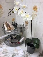 Two-tier gray metal corner caddy with miscellaneous items, placed on a kitchen countertop corner with floral tile background. Included is a 24 inch faux orchid plant with white blooms and green leaves in a black pot.