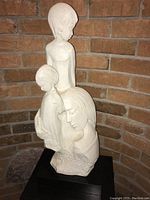 Front view of the white statue showing the mother with children with smooth facial features and flowing hair, placed against a brick background.