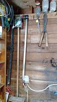 Long saw hanging vertically on wall with other tools and coiled cable on side, surrounded by wall-mounted shelves and miscellaneous items.