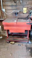 Full view of the red Kuver metal utility cart showing its general construction, flat grated top, two drawers, and wheeled base on paved floor in shed.