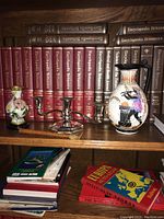 Photo showing three items on a shelf in front of Encyclopedia Britannica volumes: a floral vase on wooden stand, a silver-plated candle holder, and a decorative pitcher with painted birds and flowers.