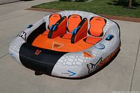 Front angled view of the 3-person inflatable water tube showing three padded seats, six handles, and bright orange and gray coloring with blue safety straps.