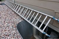 Full view of one aluminum ladder section lying on gravel next to house, showing overall length and construction.