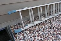 Aluminum extension ladder resting on gravel near house exterior, showing blue plastic end caps and overall wear.