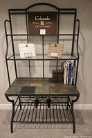 Full view of bakers rack showing metal frame, tiled work surface, glass shelves, and wine rack below with some books and display items on top