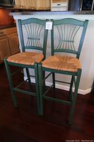 Pair of green wooden bar chairs with wicker seats, shown from the front, displaying curved top backs and vertical slats.