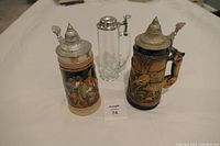 Three beer steins on white surface: two ceramic German steins, one glass stein with metal lid.