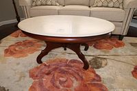 Front angle view of the coffee table showing the oval cultured marble top and mahogany wood base with curved legs on a floral rug.