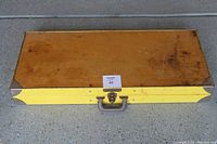 Closed rectangular yellow carrying case with metal corners and handle, showing wear marks and some discoloration.
