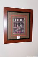 Photo of first framed picture showing Italian wine bottles and potted plants with 'CUISINE & VINS' text, warm reddish-brown wood frame with black inner border and multiple mats in brown and red shades.