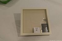 Square cream-colored shadow box on a white surface, showing front glass panel and interior backing. Small plastic bag with hardware included inside.