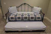 Front view of dark green metal trundle bed with upper mattress, pillows, checkered blanket, and lower pull-out mattress with fitted sheet.