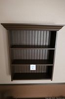 Front view of dark brown pine wood wall display shelf showing three shelves and decorative molding top with vertical paneling back