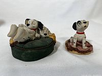 Front view of two Arden sculptures featuring Dalmatian puppies resting on a green cushion and sitting on a pink blanket.