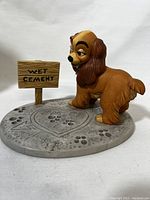 Statue of Lady from Disney's Lady and the Tramp on a cement textured base with paw prints and a sign reading 'WET CEMENT'.