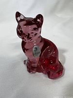 Front and side angled view of a cranberry colored glass cat figurine with a Fenton sticker on the chest. The cat is sitting upright with detailed facial features, ears, and tail. The surface is glossy and translucent.