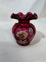 Front view showing the cranberry colored glass vase with hand painted white and yellow floral design, ruffled edge, and artist signature sticker.