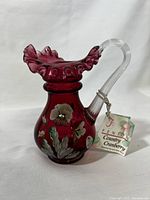 Front view of cranberry colored Fenton mini pitcher showing ruffled top edge, clear handle, hand-painted floral decoration, and attached Fenton tag.