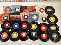 Full lot of 24 vintage 45 RPM records displayed on a flat surface. Some records have sleeves; most do not. Variety of label colors and text, showing different artists and record companies.