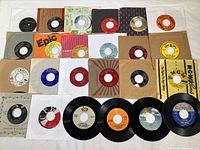 Photo showing 24 vintage 45 RPM vinyl records spread out, many with original sleeves and some without, displaying a variety of record labels and sleeve designs.