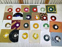 Overview of 24 vintage 45 RPM records in assorted sleeves and labels with variety of colors and branding visible.