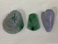 Photo showing three polished stone pendants placed side by side: carved fish design stone, smooth green pendant, and purple pinecone pendant.