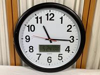 Front view of round black framed wall clock with white face, black hour and minute hands, red sweep second hand, and digital display at bottom.