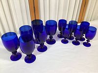Set of twelve blue glassware pieces on white surface with wooden background. Six 8-ounce goblets and six matching 4-ounce glasses visible with deep rich blue color.