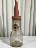 Front view of used glass motor oil bottle with plastic nozzle and label 'Mobiloil Arctic SAE 20-20 W'. Visible dirt and wear on bottle and label.
