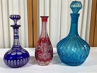 Three glass items on display: blue cut to clear decanter with stopper on left, cranberry cut to clear crystal vase in the center, and tall aqua cut to clear decanter with stopper on right.