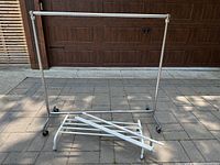 Large industrial metal clothing rack with chrome finish and caster wheels alongside disassembled white IKEA rack parts on ground.