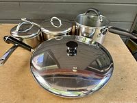 Photo of four pieces of stainless steel cookware including saucepans and frying pan with lids, all with black handles or knobs, shown on a wooden surface.