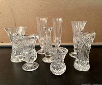 Eight assorted clear crystal bud vases grouped together, showing varied cut patterns and shapes with pedestal bases and flared rims.