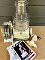 Front view of assembled food processor with bowl, blades, discs, pushers, and manuals arranged in front on table.