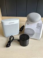 White Bose SoundLink Color II, small black Blue Piston Mini speaker, and Google Home Mini with box on display surface.