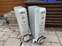 Two Delonghi Safe Heat portable standing heaters photographed upright outdoors on brick paving, showing front control panels and cords.