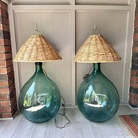 Front view of two large green glass demijohn bottles with wide bases, topped by natural woven wicker lamp shades, placed side by side on wooden floor.