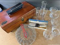 Wooden shoe shine box with metal label, wire egg basket with red handle, boxed cutlery set, and assorted champagne and sherbet glassware on table