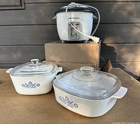 Two vintage Pyrex casserole dishes with glass lids and a Black & Decker rice cooker placed on cardboard box outdoors.