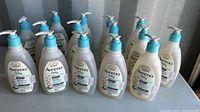 Photo showing 15 white bottles of Aveeno Baby Wash & Shampoo with blue pump tops arranged on a surface against a wall.