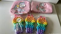 Three rainbow gradient fidget pop its shaped as cartoon characters and two pink unicorn lunch bags with printed designs, all in plastic packaging on a white surface.