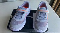 Front view of the pair of new kids Under Armour runners on the box, showing gray mesh upper with orange logo and strap.