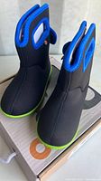Top angled view of new black kids boots with blue trim and green soles sitting on the Bogs branded box.