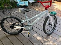 Side view of the kids bike showing white spray painted frame, black tires, black and white seat, and red handlebars. Visible damage to the back wheel area.