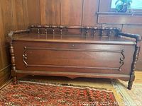 Front view of cedar chest showing carved decorative accents and turned spindle legs, positioned against wood-paneled wall.