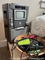 Vintage karaoke machine with front CRT screen and speaker, control buttons on side and front, two wired microphones in a wooden bowl, and multiple karaoke CDs scattered on a nearby surface.
